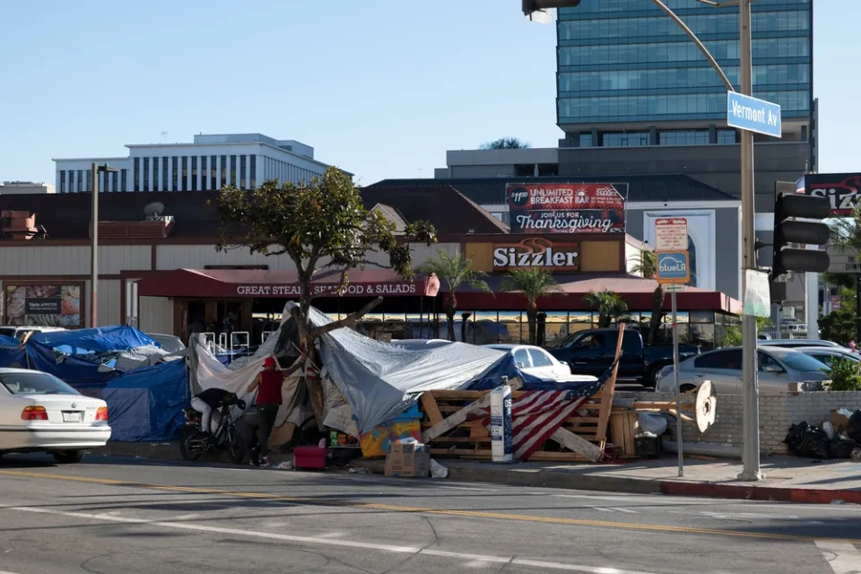 A homeless encampment fills a sidewalk in front of a Sizzler restaurant advertising Thanksgiving dinner.
